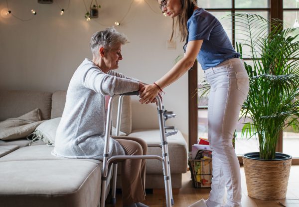 A healthcare worker or caregiver visiting senior woman indoors at home, helping her to walk.